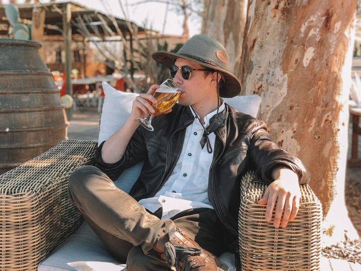 Man with a hat and shades sipping alcohol seated on a cane chair