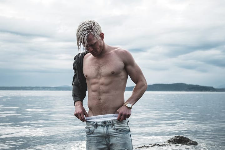 A shirtless white-haired man with a shredded physique standing by the beach