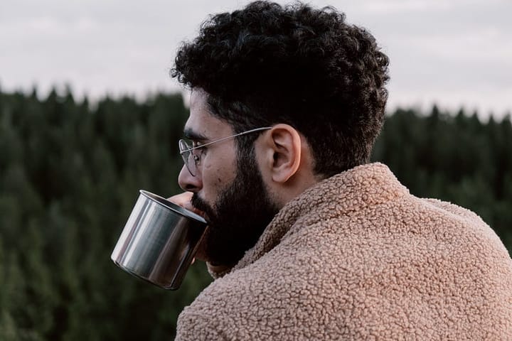Bearded and spectacled young man sipping coffee in the backdrop of a forest