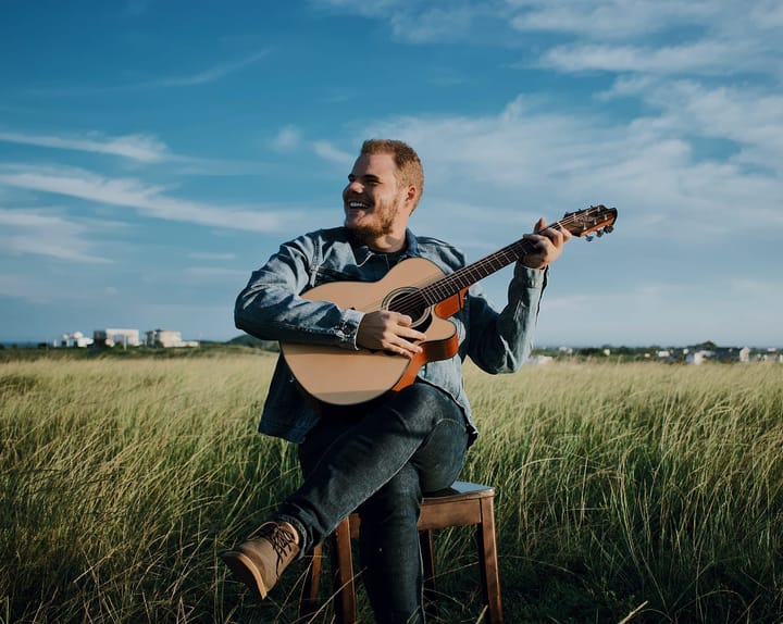 A happy man in a denim jacket playing his guitar in lush fields
