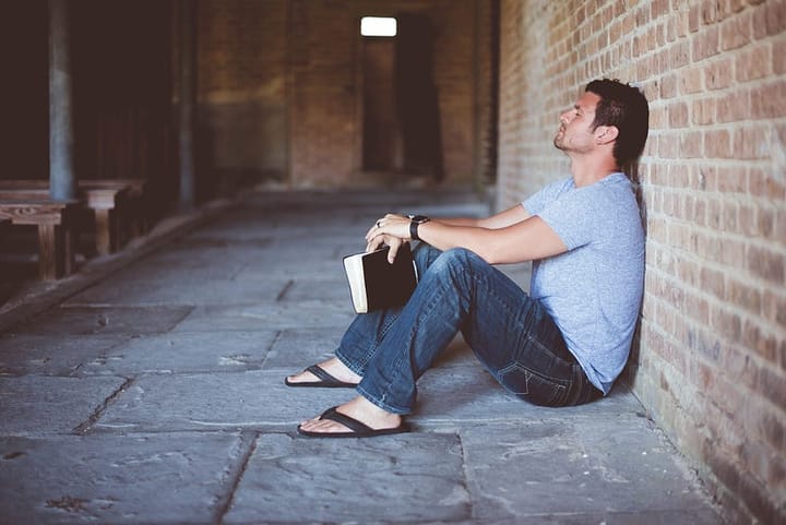 Young man sitting against a brick wall with eyes closed and a book clutched