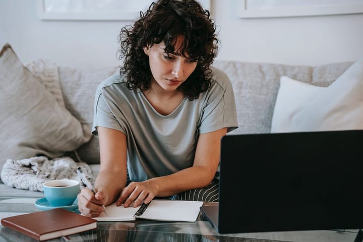 Gorgeous woman with lush black hair writing in a journal staring at a laptop screen