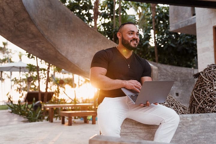 Muscular young man in a black polo and white trousers sitting with a laptop outdoors